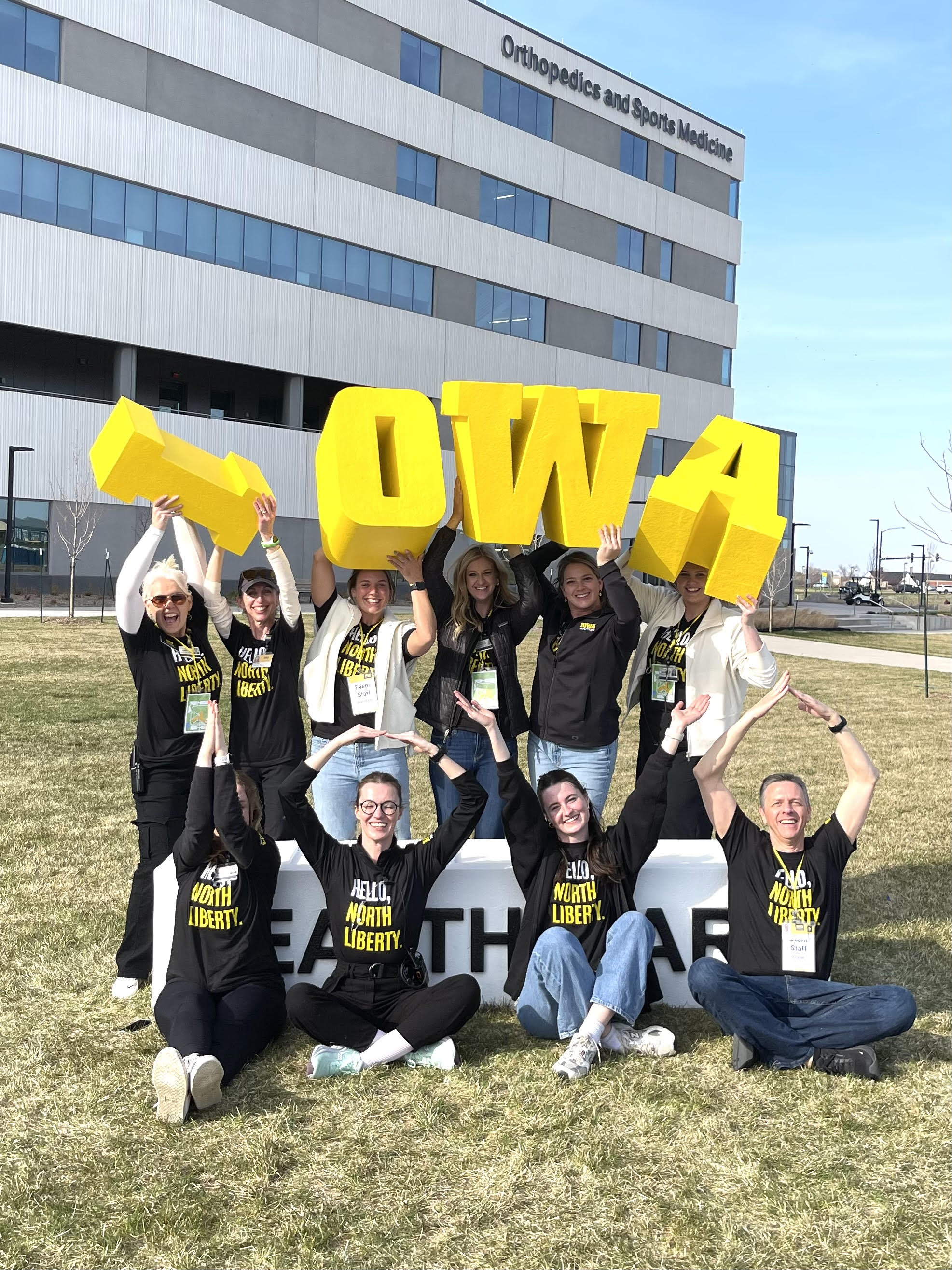 Group of smiling volunteers outside the University of Iowa Hospitals and Clinics holding large yellow block letters spelling 'IOWA,' wearing matching black 'Hello North Liberty' t-shirts for a bulk hospital project.